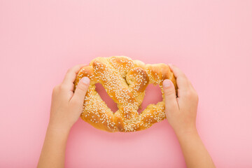 Little girl hands holding soft pretzel with sesame seeds on light pink table background. Pastel color. Closeup. Point of view shot. Top down view.