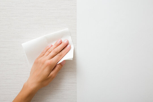 Young adult woman hand holding white dry paper napkin and wiping surface from glue after light gray wallpaper gluing. Closeup. Front view. Repair work of home.