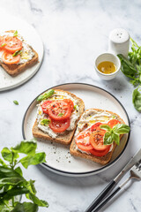 Sandwiches or toasts with tomatoes, cream cheese, olive oil and basil on a plate on white marble background. Traditional italian mediterranean food