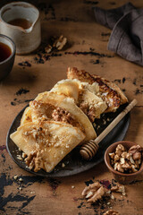 Crepes or thin pancakes with honey and nuts on brown plate over wooden background. Delicious homemade crepes for breakfast dark moody
