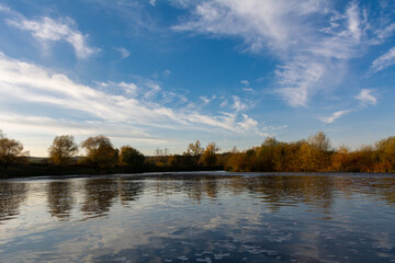 Amazing autumn landscape with calm river, blue sky with clouds and trees
