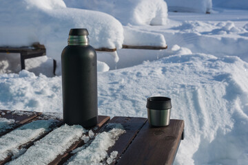 A vacuum travel thermos standing on a wooden table in the open air, and next to a Thermos mug with...