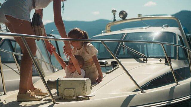 Mother pulls out little boy of boat hatch with girl in sunny city port on summer day. Cute children with woman leave moored yacht against mountains