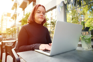 Portrait of a young student girl working remotely from laptop while sitting at a table in a cafe. Female student studying at park using modern laptop at bright sunny day. Working outdoor.