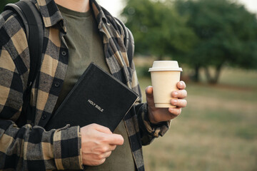 Bible and paper cup in the hands of a Christian man outside on a blurred background of nature.