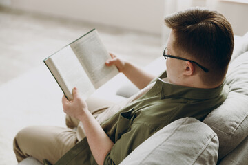 Side view young calm man with down syndrome wear glasses casual clothes reading book sits on grey...