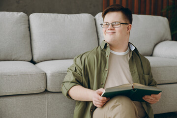 Young man with down syndrome wearing glasses casual clothes reading book look aside sits near grey sofa couch stay at home flat rest spend free time in living room. Genetic disease world day concept.