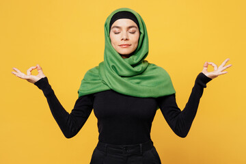 Young muslim woman wear green hijab abaya black clothes spread hands in yoga om aum gesture relax meditate try calm down isolated on plain yellow background. People uae middle eastern islam concept.
