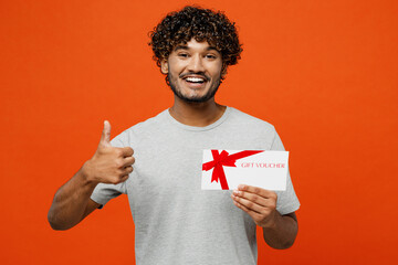 Young fun happy Indian man he wears t-shirt casual clothes hold gift certificate coupon voucher card for store show thumb up isolated on orange red color background studio portrait. Lifestyle concept. - obrazy, fototapety, plakaty