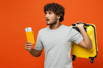 Traveler sad Indian man wear t-shirt casual clothes hold suitcase passport ticket isolated on plain orange background. Tourist travel abroad in free time rest getaway. Air flight trip journey concept.