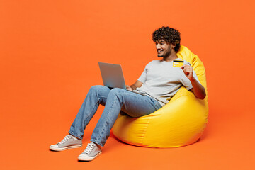 Full body young Indian man wear t-shirt casual clothes sit in bag chair use laptop pc computer hold...