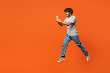 Fototapeta na wymiar Full body side view young smiling happy Indian man wears t-shirt casual clothes jump high hold in hand use mobile cell phone isolated on orange red color background studio portrait. Lifestyle concept.