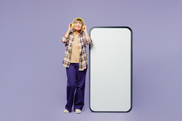 Full body elderly woman 50s years old wear beige t-shirt shirt casual clothes headphones big huge blank screen mobile cell phone smartphone with area listen music isolated on plain purple background.