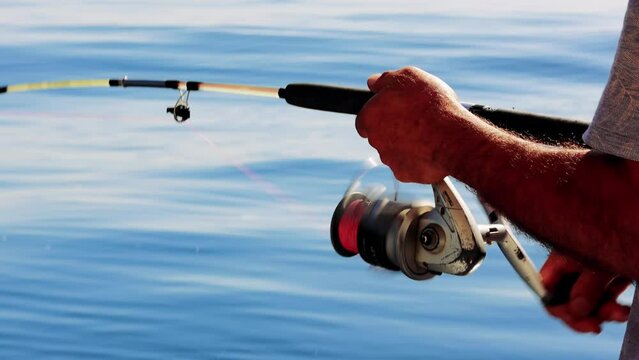 The hands of an adult male fisherman close-up spin a reel on a spinning rod while fishing on a sunny summer day from the side of a sea boat against the backdrop of a clear blue sea