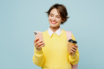 Young woman student wearing casual clothes sweater backpack bag hold takeaway delivery cup coffee to go use mobile cell phone isolated on plain blue background. High school university college concept.