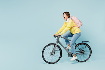 Full body side view young shocked surprised woman student wears casual clothes sweater backpack bag ride bicycle look aside isolated on plain blue background. High school university college concept.