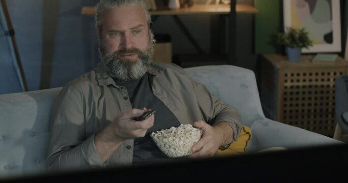 Mature man turning on TV with remote control choosing channel holding popcorn watching program in evening in living room. Mass media and leisure activity concept.
