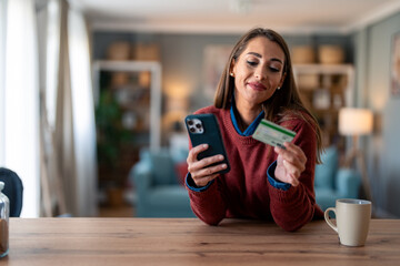 Happy young woman customer shopper holding credit card using cell phone mobile app paying online making purchase in e-commerce digital store on smartphone at home. - obrazy, fototapety, plakaty