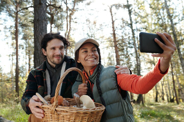 Young intercultural dates with basket of mushrooms looking at smartphone camera with smiles during selfie in autumn forest