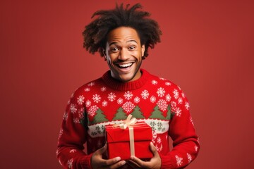 Fototapeta premium Happy black man wearing in ugly Christmas sweater with red gift on red background. Ugly sweater Day on December.