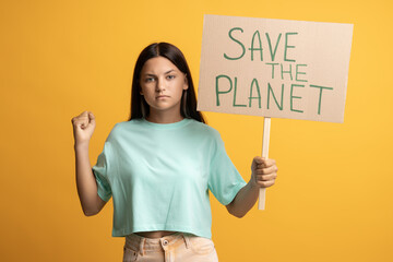 Serious confident brunette teen girl eco activist showing holding save the planet poster on yellow...