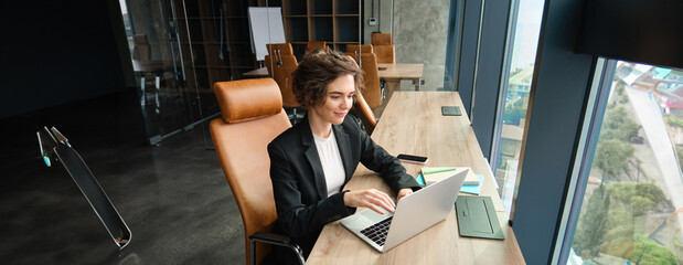 Portrait of corporate woman in suit, working in office, using laptop, typing, does her job in coworking space - obrazy, fototapety, plakaty
