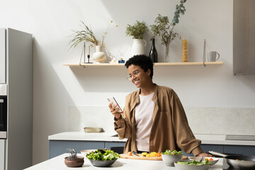 Happy cheerful African chef girl making video call on smartphone from home kitchen, cooking healthy meal for dinner, using mobile phone at table with dish, fresh vegetables, organic food ingredient
