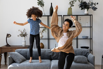 Cheerful excited young African mom teaching pretty preschool daughter kid to dance in living room,...