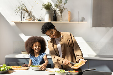 Happy carefree young Black mom and daughter kid cooking healthy vegetarian meal for family dinner in home kitchen, slicing, frying fresh vegetables, talking, laughing, chatting, enjoying leisure - obrazy, fototapety, plakaty