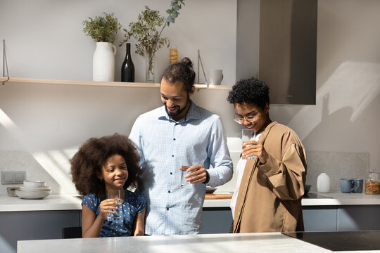 Happy millennial Black parents and little daughter kid keeping healthy nutrition, drinking clean natural water in kitchen, using transparent glasses, talking, smiling, laughing. Family healthcare