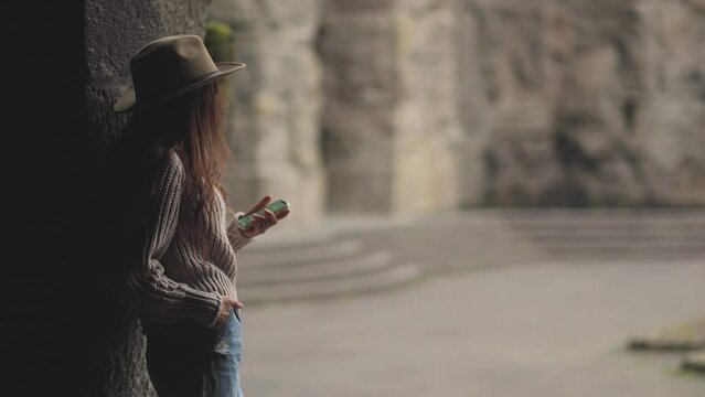 A woman in a hat types a message on her phone, waiting for a meeting, gazing into the distance. Beautiful style and a tourist spirit. A meeting on a weekend. A cinematic atmosphere. Shadow silhouette