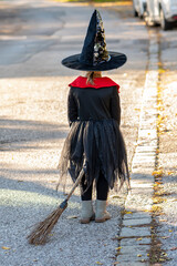 Girl in black witch costume with broom on Halloween walking down the street, back view from behind.