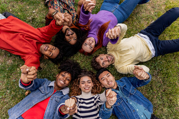 A diverse group of friends are lying on a grass in a circle with their heads together and hands in...