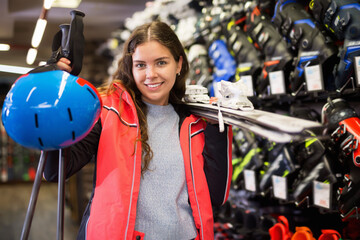Portrait of a positive woman with brown hair choosing colorful skis, boots and helmet in sports store