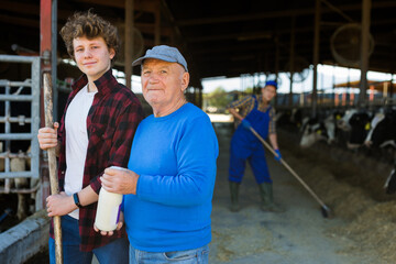Fototapeta na wymiar Elderly farmer and his assistant grandson with bottle of milk on the background of cows in a stall