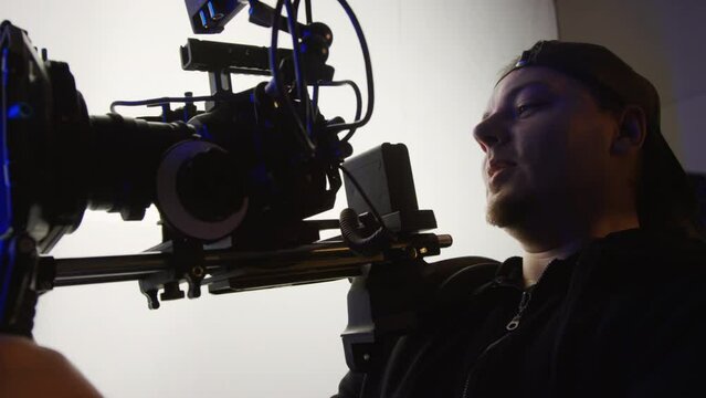 Medium shot of Caucasian cameraman in baseball cap holding professional video camera on shoulder rig, adjusting frame and chatting to production crew, while working backstage on commercial shoot