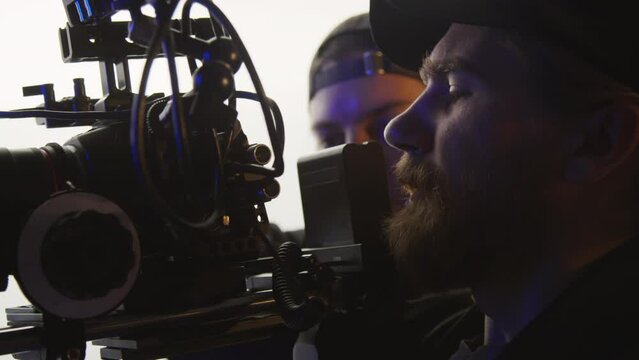Close-up shot of faces of male videographer shooting footage on professional video camera or camcorder on shoulder rig and looking into viewfinder, assistant standing and watching