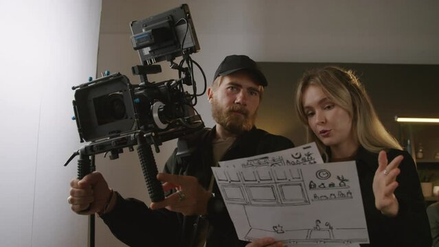 Medium shot of young blonde female director standing in studio with storyboard sheets, talking to cameraman, describing scenes and pointing, and videographer with professional camera listening