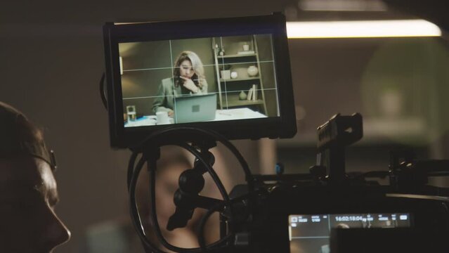 Close-up shot of male videographer in baseball cap setting up professional video camera with additional screen, connecting data cables, and female presenter with laptop getting ready for shoot