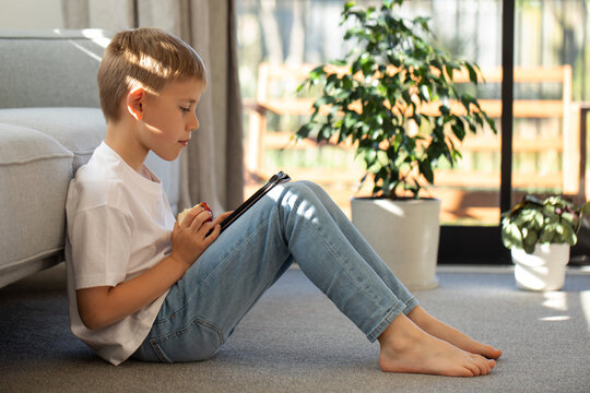 A cute schoolboy is sitting on the floor in the living room and reading an e-book. E-book. - Powered by Adobe