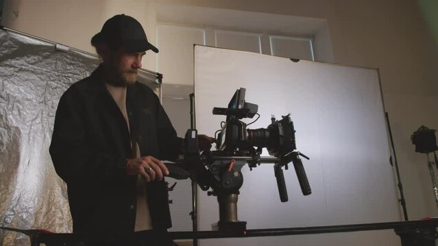 Medium shot of Caucasian male videographer in black shirt and baseball cap standing in studio before shoot, adjusting and testing his camera, then showing thumb up