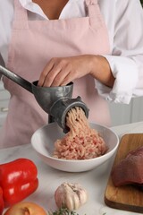 Woman making chicken mince with metal meat grinder at white table in kitchen, closeup