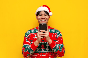 young asian man in christmas sweater and santa hat using smartphone on yellow isolated background