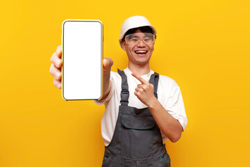 Asian guy builder in uniform and safety glasses shows blank smartphone screen on yellow isolated...