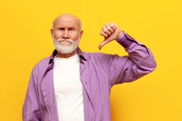 dissatisfied old bald grandfather with gray beard in purple shirt shows dislike on yellow isolated...