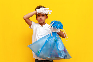 african american boy teenager volunteer in white t-shirt and gloves removes trash and holds trash bag