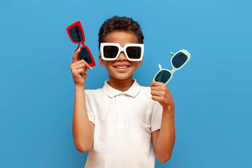 african american teenage boy with glasses holding different sunglasses on blue isolated background