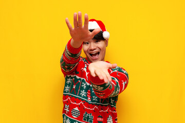 young asian man in christmas sweater and santa hat shows hands forward on yellow isolated background