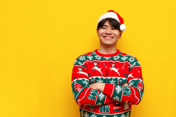 young asian man in christmas sweater and santa hat standing with crossed arms over yellow isolated...