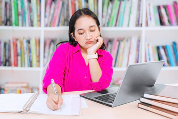 Exhausted and bored female student studying on the desk with laptop and books with bookshelf background